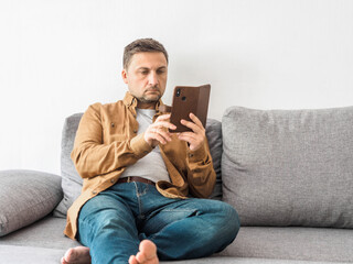 Middle aged man in beige shirt and jeans sits at home on gray sofa and looks intently at the phone. Smartphone in hand. Blogging, online education or remote work concept