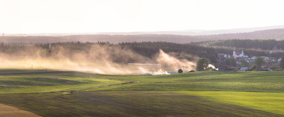 Fototapeta premium Panorama of the countryside of Belarus. Agricultural fields view. Fresh green field and a small European town. Smoke from burnt grass spreads across the field. Large forest in fog on the background
