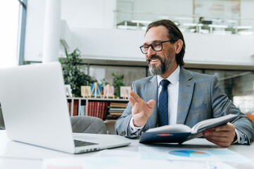 Smiling male employee wearing in formal suit, using laptop, makes notes at online conference.Mature successful businessman sits at the desk, working remotely, communicate with colleagues by video call