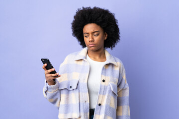 Young African American woman using mobile phone isolated on purple background with sad expression
