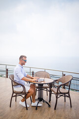 Obraz premium Handsome successful young male businessman sitting at a table by the pool with a laptop overlooking the Mediterranean Sea. Remote work on vacation. Vacation concept