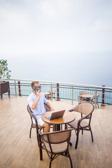 Handsome successful young male businessman sitting at a table by the pool with a laptop overlooking the Mediterranean Sea. Remote work on vacation. Vacation concept