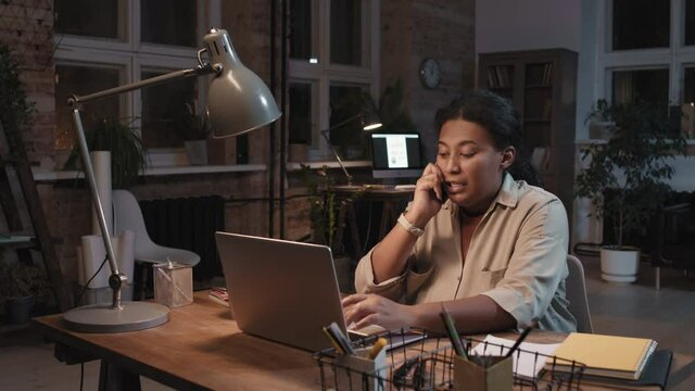 Medium slow-motion arc shot of hard-working african american woman talking on phone while working on laptop in dark loft office alone at night