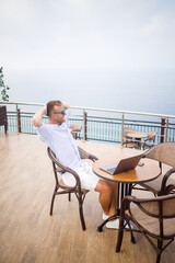 Handsome successful young male businessman sitting at a table by the pool with a laptop overlooking the Mediterranean Sea. Remote work on vacation. Vacation concept