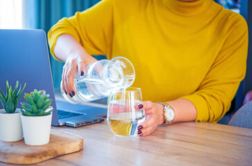 A young business woman sitting at working desk pouring drinking water into the glass. In the idea of drinking more water in the day to substitute the hungriness to losing weight and be more healthy. 