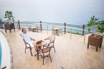 Handsome successful young male businessman sitting at a table by the pool with a laptop overlooking the Mediterranean Sea. Remote work on vacation. Vacation concept