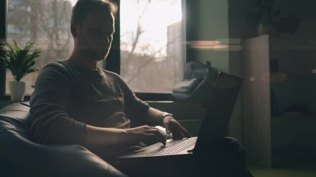 A handsome man is looking on monitor and typing on keyboard at house. A casual programmer is using laptop.
