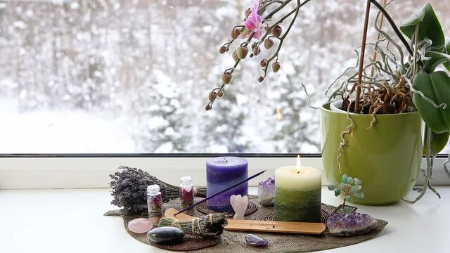 Small good feng shui altar in home on window sill on leaf shape table mat, snowy Nordic nature on background. Incense candle smoking, gemstones and orchid flower pot.