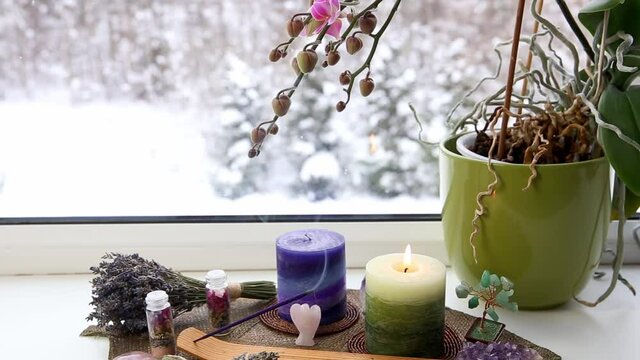 Small good feng shui altar in home on window sill on leaf shape table mat, snowy Nordic nature on background. Incense candle smoking, gemstones and orchid flower pot.