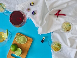 Various glasses of wine and spirits on white crumpled cloth with blue background, table with cut lime slices, overhead view