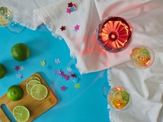 Various glasses of wine and spirits on white crumpled cloth with blue background, table with cut lime slices and remnants of confetti and party, overhead view