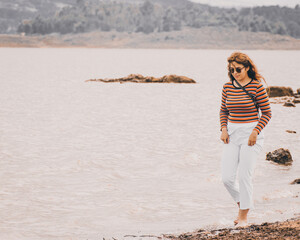 Chica joven y bonita vestida con una blusa naranja y un pantalón blanco es turista en una playa pequeña, esta sosteniendo su cámara mientras camina por la orilla haciendo fotos
