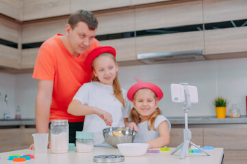 Father teaches two daughters to cook dough in the kitchen. The family is filming a culinary video on a smartphone.