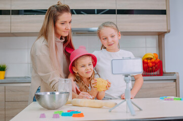 Mom teaches her daughters to cook dough in the kitchen. The family is filming a culinary video on a smartphone.