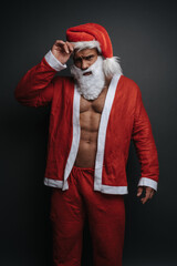 Frowning muscular man in Santa Claus costume in studio. Emotional portrait of handsome male model in Christmas outfit at grey background.