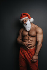Shirtless muscular man in Santa hat with holding his right hand on his stomach. Sexy Christmas hunk in studio at grey background.