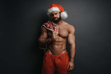 Sad Santa Claus with red plaid in studio. Sexy muscular guy in Santa hat at grey background. Shirtless man in Christmas pants holding  checkered blanket on his chest.