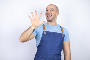 Young bald man wearing apron uniform over isolated white background showing and pointing up with fingers number five while smiling confident and happy