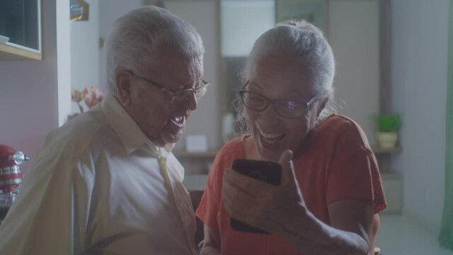 An Elderly Couple In The Home Kitchen Talking On The Cell Phone With Friends Or Relatives Using Facetime Video Calling Application.