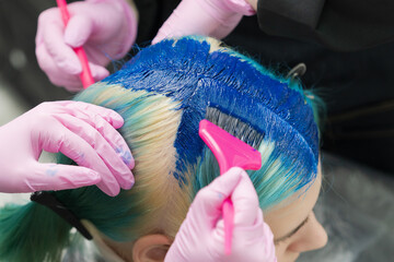 Two hairdressers using pink brush while applying blue paint to female customer with emerald hair...