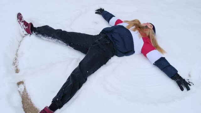 Cheerful young woman making angel lying on snow and moving hands like wings, pretending to fly, resting and enjoying winter, remembering childhood, winter magic, slow motion