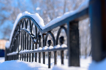 A snow-covered black forged fence made of thick metal profiles is positioned diagonally in the...