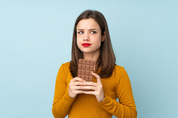 Teenager girl isolated on blue background taking a chocolate tablet and having doubts