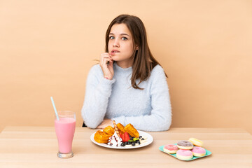 Teenager girl eating waffles isolated on beige background with confuse face expression