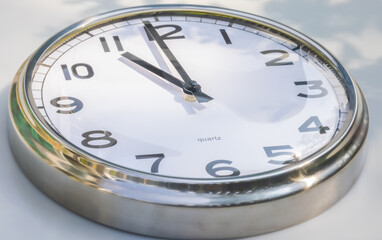 A Modern white aluminum wall clock in sunlight with shadows on the  clock face. Close up to a wall clock. Reflections in the aluminum frame. The pointers at 11 o'clock