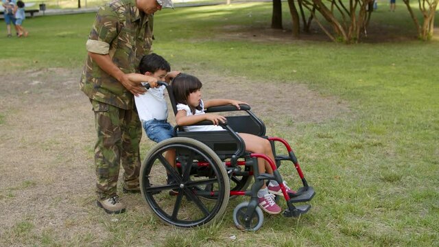 Military father walking with son and disabled daughter in park, wheeling wheelchair with girl on grass. Dolly shot. Parenthood or disability concept