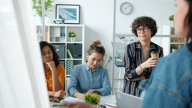 Serious office workers are listening to mature woman making presentation with whiteboard in workplace. Businesspeople and ambitions women concept.