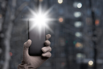Gloved hand holds a smartphone with a burning flashlight with lighting windows of Moscow dwelling...