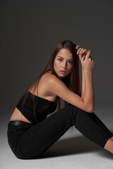 Studio portrait of young slim tanned caucasian girl in black jeans and bando top sitting and posing against grey studio background