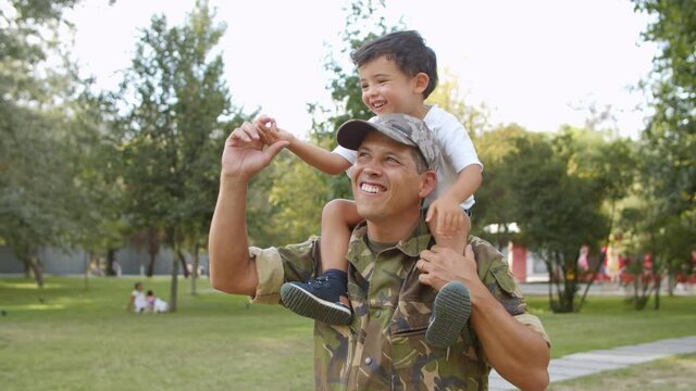 Happy cute little boy riding dads shoulders in park. Military daddy piggybacking son outdoors, walking in park. Dolly shot. Soldier father or fatherhood concept