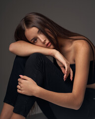 Full length studio portrait of young slim tanned caucasian girl in black jeans and bando top sitting and posing against grey studio background