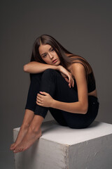 Fototapeta na wymiar Full length studio portrait of young slim tanned caucasian girl in black jeans and bando top sitting and posing against grey studio background
