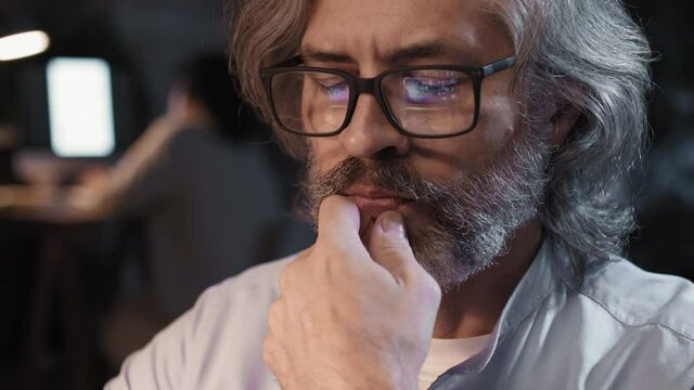 Slow-motion close-up of thoughtful senior office worker sitting at desk at night working