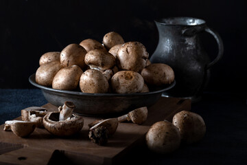 Fresh champignon (button) mushrooms in bowl on a wooden board front of the dark background.
