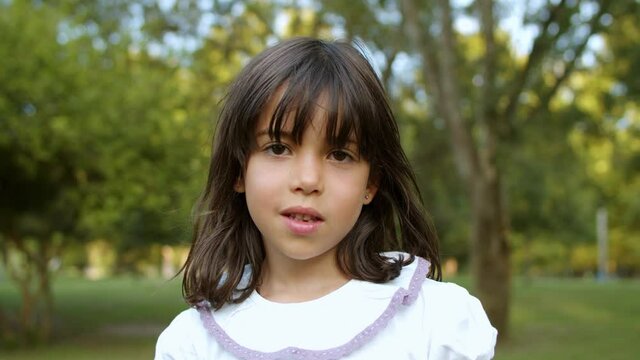 Pensive cute little black haired girl standing in city park, looking at camera and smiling. Kid enjoying leisure time outdoors in summer. Headshot, closeup. Childhood concept