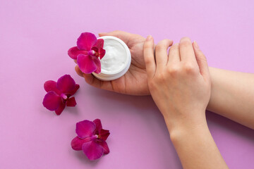 Cosmetic background with hands, soft white cream and fragile magenta colored orchids.Moisturizing facial cream in a jar and blooming orchid flowers on pink background, skin care cosmetics.