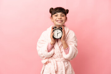 Little caucasian girl isolated on pink background in pajamas and holding clock with happy expression