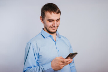 A guy in a blue shirt holds a smartphone on a white background. A male office worker is typing text on the phone. Photo in studio