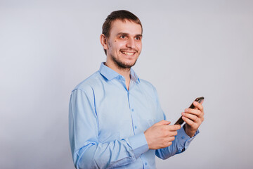 A guy in a blue shirt holds a smartphone on a white background. A male office worker is typing text on the phone. Photo in studio