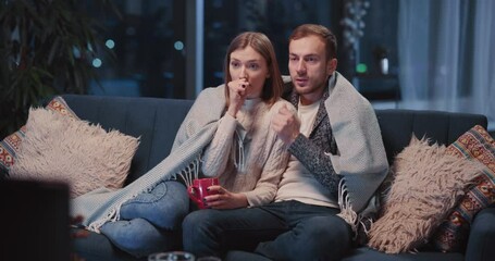 Emotional caucasian man and woman couple watching TV horror movie. Young girl getting scared hugging tight her boyfriend. Emotions. Excitement. - Powered by Adobe