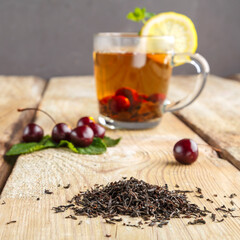 black tea in a glass cup with mint cherries and lemon on a wooden table next to fresh cherries and sprinkled tea leaves.