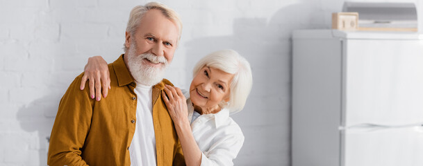 Smiling elderly woman hugging husband in kitchen, banner