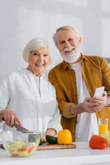 Smiling senior couple with smartphone looking at camera while cooking on blurred foreground