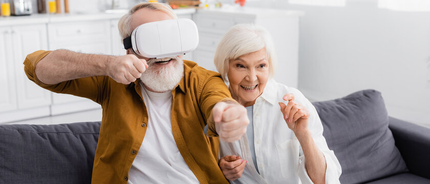 Smiling senior woman sitting near husband in vr on couch, banner - Powered by Adobe