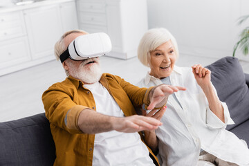 Senior man using vr headset near smiling wife in living room