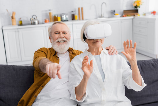 Cheerful senior man pointing with finger on blurred foreground near wife in vr headset